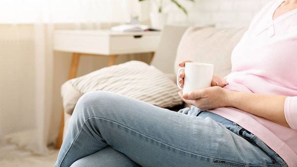 Un primer plano de mujer sentada cómodamente en un sofá beige, sosteniendo casualmente una taza blanca en su mano, con la luz del sol filtrándose a través de la habitación, recortada - Foto, imagen