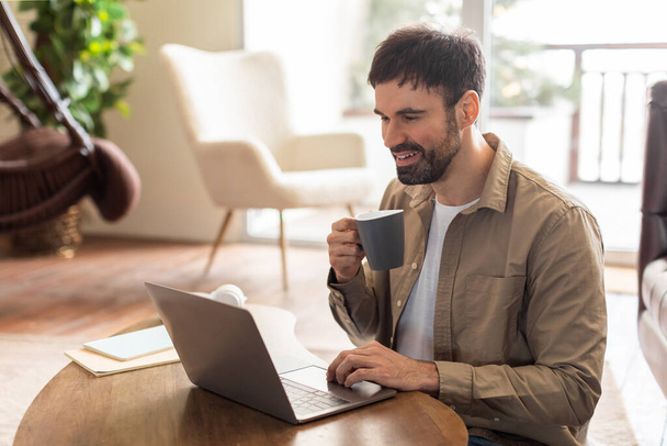 Een man in een nonchalante kledij zit aan een tafel en concentreert zich op zijn laptop scherm. Naast hem, is er een kopje koffie, suggereert een werkomgeving - Foto, afbeelding