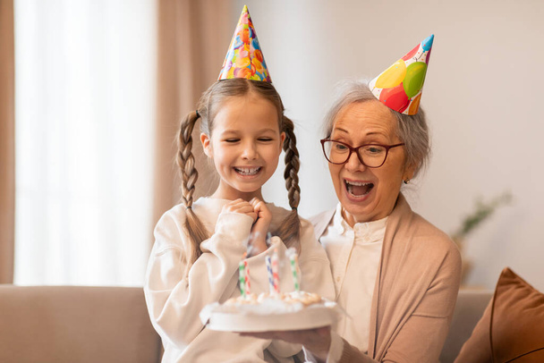 Літня жінка і молода дівчина носять різнокольорові капелюхи на день народження в кімнаті, прикрашеній для святкування. Вони посміхаються і тримають повітряні кулі в оточенні подарунків і вечірок прикрас. - Фото, зображення