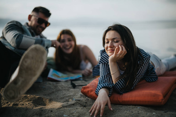 Τρεις φίλοι διασκεδάζουν σε μια αμμώδη παραλία, μια γυναίκα ξαπλώνει ενώ ένα ζευγάρι κάθεται δίπλα της, γελώντας και απολαμβάνοντας την ήρεμη παραθαλάσσια ατμόσφαιρα. - Φωτογραφία, εικόνα