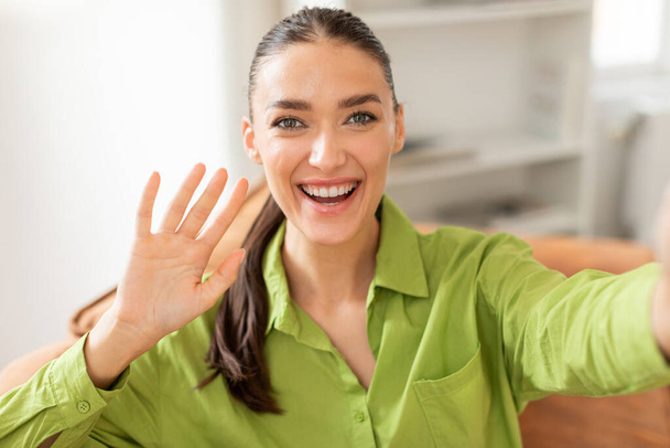 Μια χαρούμενη νεαρή γυναίκα με μακριά μαλλιά δεμένα πίσω απλώνει το χέρι της σε ένα κύμα, συμμετέχοντας σε μια περιστασιακή βιντεοκλήση, ενώ ντύνεται σε μια ζωντανή πράσινη μπλούζα - Φωτογραφία, εικόνα