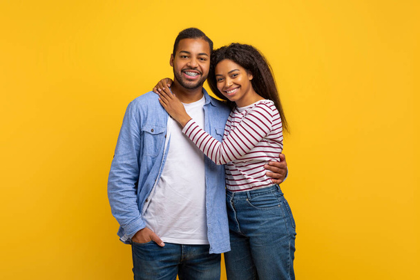 Casal afro-americano de pé lado a lado na frente de um fundo amarelo brilhante. Eles estão olhando diretamente para a câmera, ambos vestindo roupas casuais, abraçando - Foto, Imagem