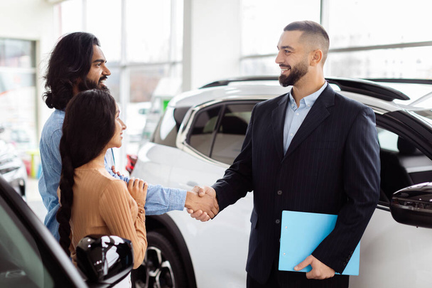 Продавець автомобілів у костюмі розширює рукостискання усміхненому індійцю, жінка стоїть поруч з ним, можливо, завершує продаж або вітає їх всередині автосалону - Фото, зображення