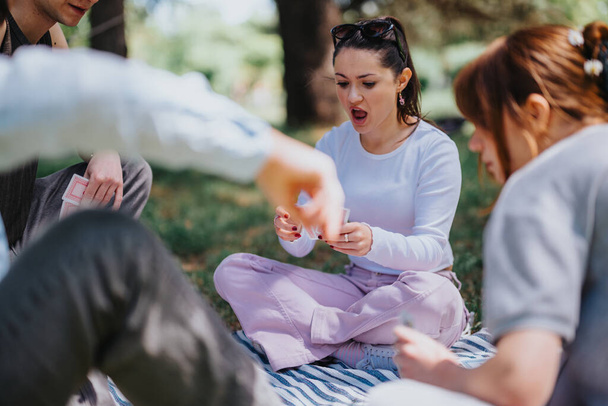 Un grupo de amigos disfruta de un juego de cartas casual al aire libre en una tarde soleada, compartiendo momentos de emoción y compromiso. - Foto, Imagen
