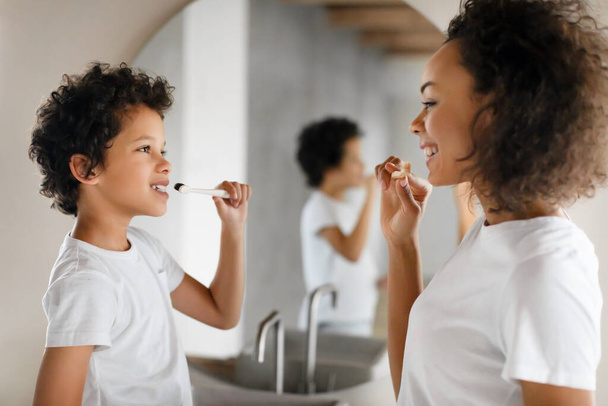 Afroamerykanka myje zęby obok małego chłopca. Stoją przed umywalką w łazience. Kobieta trzyma szczoteczkę, podczas gdy mały chłopiec patrzy uważnie.. - Zdjęcie, obraz