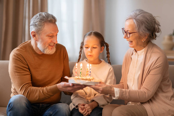 Una giovane ragazza seduta tra i suoi nonni sorridenti sta spegnendo le candele su una torta di compleanno che suo nonno sta tenendo in mano. Sembrano seduti comodamente in un soggiorno - Foto, immagini