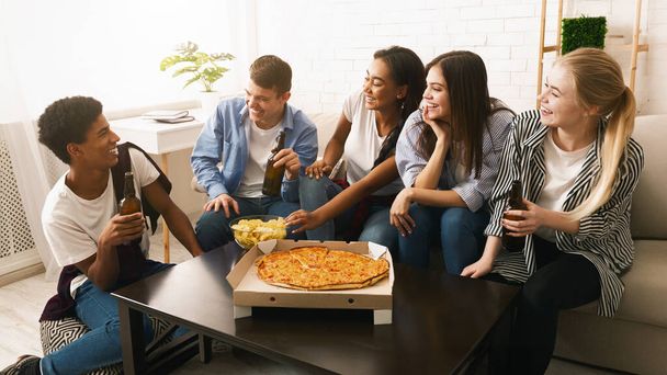Egy csapat tinédzser ül az asztal körül, és pizzát esznek. A barátok beszélgetnek, miközben a sajtos és sós ételeket eszik, alkalmi és társadalmi légkört teremtve.. - Fotó, kép