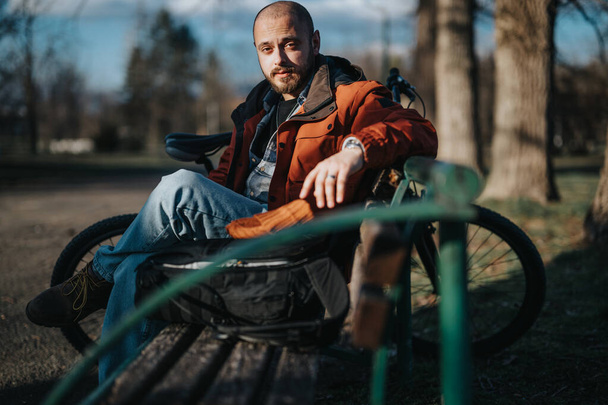 Розслаблений молодий чоловік у повсякденному одязі сидить зі своїм мотоциклом у безтурботній обстановці парку, дивлячись споглядальний. - Фото, зображення