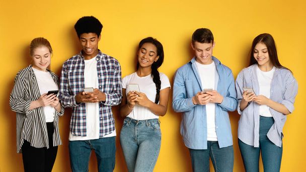 A diverse group of five multiethnic teenagers stand side-by-side against a vibrant yellow backdrop, each engrossed in their own smartphone - Photo, Image