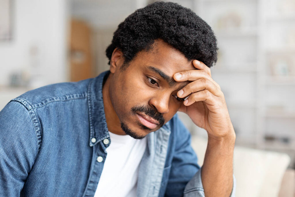 Upset African American man is seated on a couch, with his hand resting on his head in a thoughtful gesture. He appears deep in contemplation or possibly experiencing stress or fatigue - Photo, Image