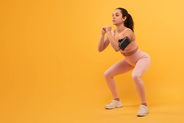 Egy fiatal nőt, aktív ruhába öltözve, mozgásban rögzítenek, miközben guggoló gyakorlatot végez, koncentrál és jó formában tartja a karjait maga előtt tartva, edz sárga háttér mellett. - Fotó, kép