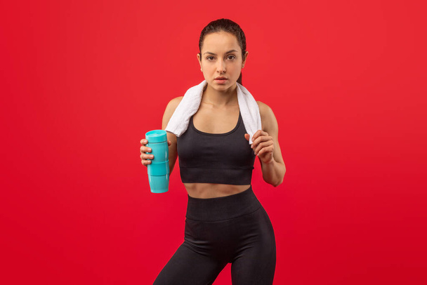 Uma mulher focada e atlética vestindo uma roupa esportiva preta e uma toalha branca ao redor do pescoço está contra um fundo vermelho vívido, segurando uma garrafa de água azul, provavelmente fazendo uma pausa - Foto, Imagem
