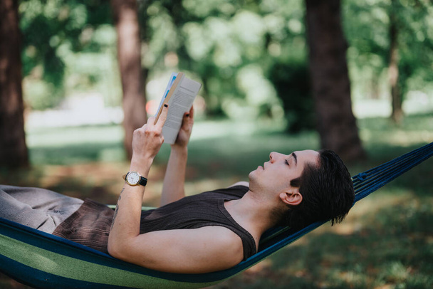 A young adult male relaxes with a book in a hammock, set against a lush green park background on a sunny day. Concept of leisure and relaxation. - Photo, Image