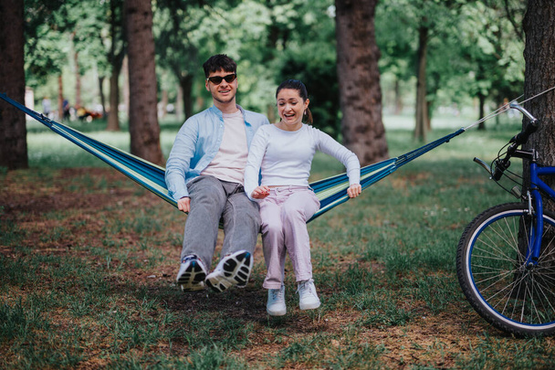 Ein junges Paar genießt einen verspielten Moment auf einer blauen Hängematte in einem üppig grünen Park, neben sich parkt sein Fahrrad, das Freizeit und Glück verkörpert. - Foto, Bild