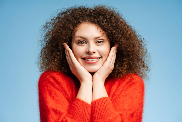 Ritratto di donna allegra sorridente con i capelli ricci che indossa elegante maglione rosso guardando la fotocamera in piedi isolata su sfondo blu. Concetto di shopping, bellezza naturale, cura dentale - Foto, immagini