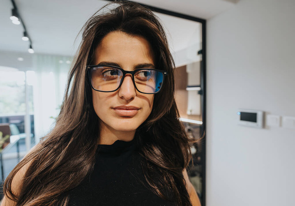 Közelkép egy magabiztos, elegáns szemüveges fiatal nőről, modern irodai háttérrel, professzionalizmussal és intelligenciával. - Fotó, kép