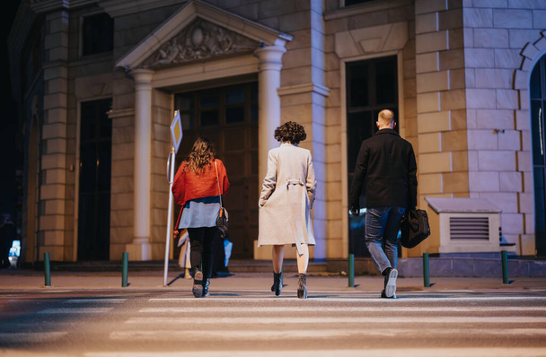 Drie vrienden doorkruisen een zebrapad in een stedelijke omgeving onder de avondhemel. Het begrip stadsleven en nachtelijke vrijetijdsbesteding vastleggen. - Foto, afbeelding