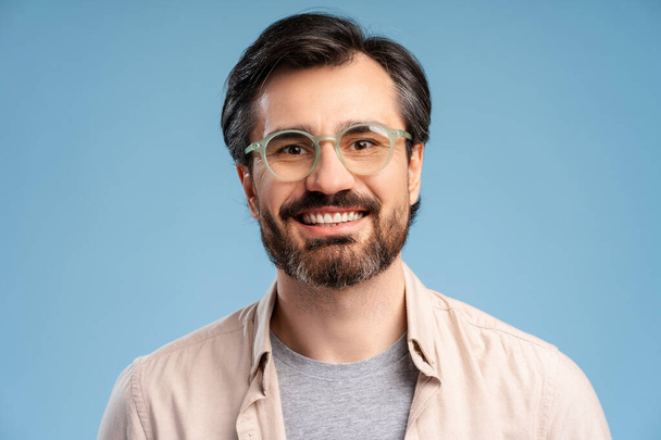 Портрет щасливого бородатого чоловіка в окулярах позує в студії, дивлячись на камеру, ізольовану на синьому фоні. Покупки, концепція продажу. Концепція реклами - Фото, зображення