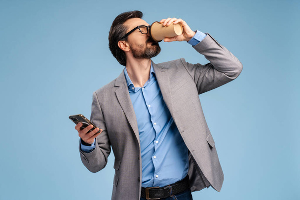 Portrait of bearded young man in jacket and eyeglasses enjoying coffee drink and holding smartphone, posing against blue background. Technology, business concept - Photo, Image