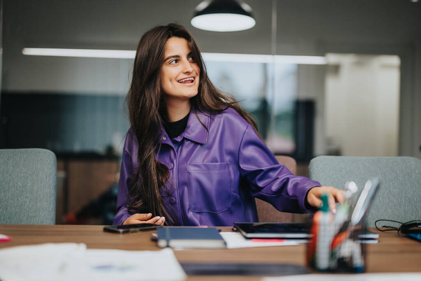 Una giovane donna concentrata che indossa una giacca viola vibrante gode il suo lavoro in un ambiente di ufficio vivace, mostrando produttività ed energia positiva sul posto di lavoro. - Foto, immagini
