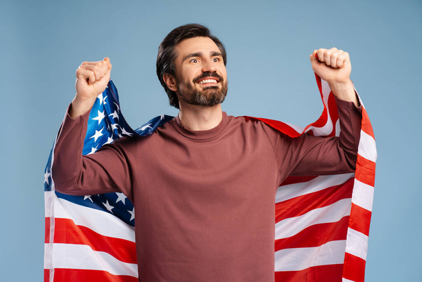 Muotokuva hymyilevä nuori komea mies Amerikan lippu poseeraa studiossa, juhlii, eristetty sininen. Memorial Day juhla, itsenäisyyspäivä - Valokuva, kuva