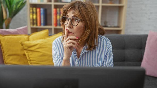 Egy töprengő középkorú nő szemüveggel, aki otthon ül a kanapén és tévét néz.. - Fotó, kép