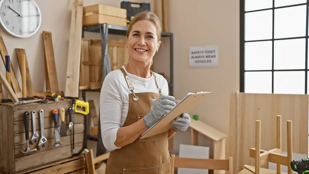 Una donna matura sorridente in un laboratorio di falegnameria che prende appunti su un blocco appunti circondato da attrezzi per la lavorazione del legno e progetti di mobili in legno. - Foto, immagini