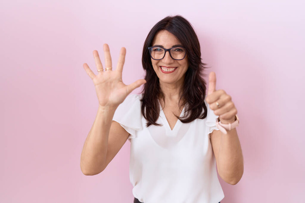 Mujer hispana de mediana edad vistiendo camiseta blanca casual y gafas que muestran y señalan con los dedos número seis mientras sonríe confiada y feliz.  - Foto, imagen