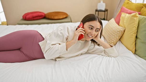 Sorridente giovane donna ispanica sdraiata sul letto a parlare su smartphone in una luminosa camera da letto con cuscini colorati. - Foto, immagini