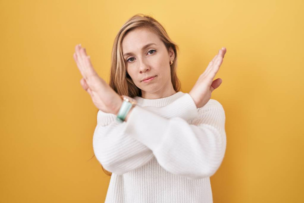 Jeune femme caucasienne portant un pull blanc sur fond jaune expression de rejet croisant les bras faisant signe négatif, visage en colère  - Photo, image