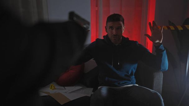 Um oficial apontando uma arma para um homem com as mãos levantadas em uma sala mal iluminada com cortinas vermelhas e marcadores de evidência. - Foto, Imagem