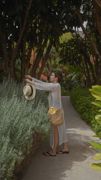 Молода жінка досліджує пишну зелень в бальзамі на відкритому повітрі, випадково тримаючи її солом'яний капелюх і кошик. - Фото, зображення