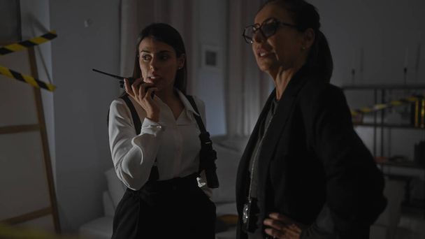 Two professional women investigators discuss a case indoors with one speaking into a walkie-talkie. - Photo, Image