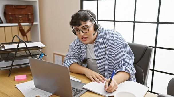 Ένας bespectacled άνθρωπος με ακουστικά εργάζεται εντατικά σε ένα φορητό υπολογιστή σε ένα σύγχρονο γραφείο, γράφοντας σημειώσεις. - Φωτογραφία, εικόνα
