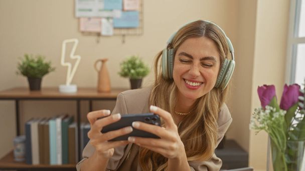 Smiling young caucasian woman wearing headphones plays a mobile game at home. - Photo, Image