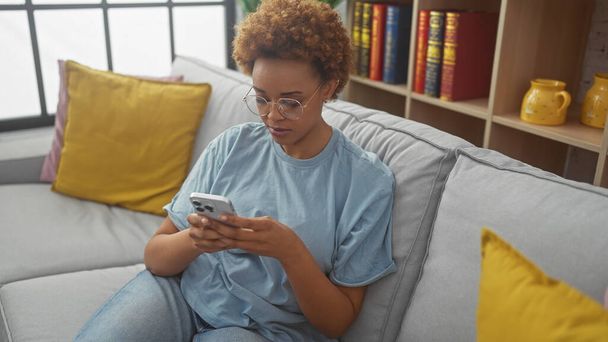 Una atractiva mujer afroamericana usando un teléfono inteligente mientras se relaja en un cómodo sofá interior. - Foto, imagen
