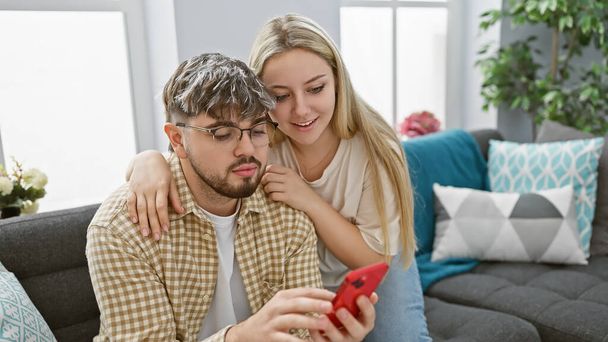 Un hombre y una mujer acurrucados en un sofá en una acogedora sala de estar, mirando un teléfono inteligente juntos, irradiando amor y vibraciones familiares. - Foto, Imagen