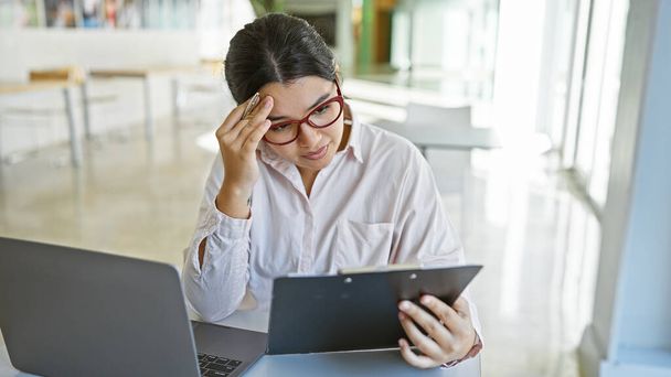 Eine junge hispanische Frau schaut besorgt, während sie ein Tablet in der Hand hält und an einem Schreibtisch im Büro sitzt, mit einem Laptop und einem lichtdurchfluteten Raum im Hintergrund.. - Foto, Bild