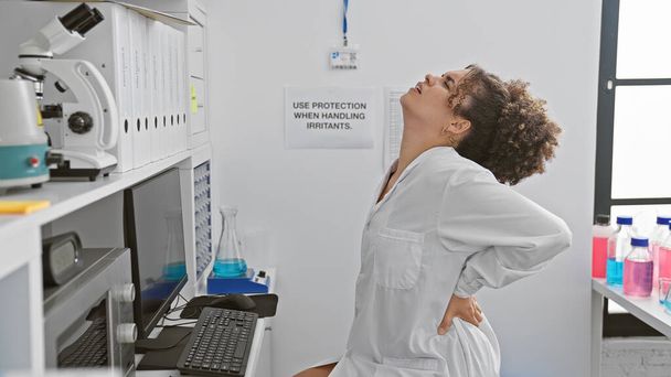 Una giovane donna con i capelli ricci che indossa un camice bianco si allunga la schiena in un ambiente di laboratorio al coperto. - Foto, immagini