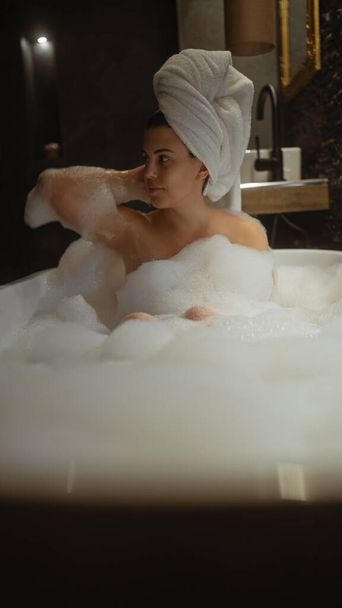 Una mujer joven disfruta de un lujoso baño de espuma en un ambiente de baño en casa bien equipado. - Foto, imagen