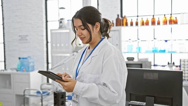Młoda Latynoska kobieta w białym fartuchu laboratoryjnym z tabletką w nowoczesnym otoczeniu laboratoryjnym, skupiona i profesjonalna. - Zdjęcie, obraz