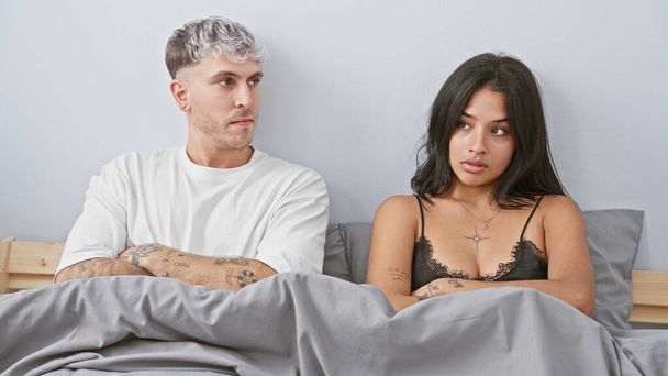 Ένας άνδρας με τατουάζ και μια γυναίκα με ένα κολιέ κάθονται χωριστά στο κρεβάτι, υπονοώντας ένταση στη σχέση τους μέσα σε ένα σκηνικό κρεβατοκάμαρας. - Φωτογραφία, εικόνα