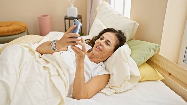 成熟したヒスパニック女性は,ベッドで快適に横たわりながらスマートフォンを使用して寝室でリラックスし,朝のルーチンに浸ります. - 写真・画像