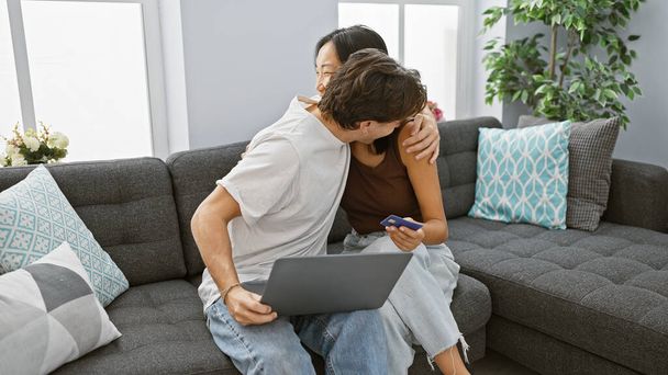 Interracial couple embracing in living room with laptop and credit card, signifying online shopping or financial planning. - Zdjęcie, obraz