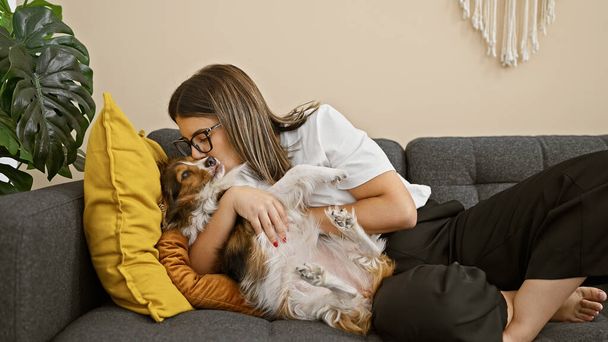 Hispán nő szeretettel ölelkezik a bolyhos kutya egy hangulatos szürke kanapén beltéri körül élénk lakberendezési. - Fotó, kép