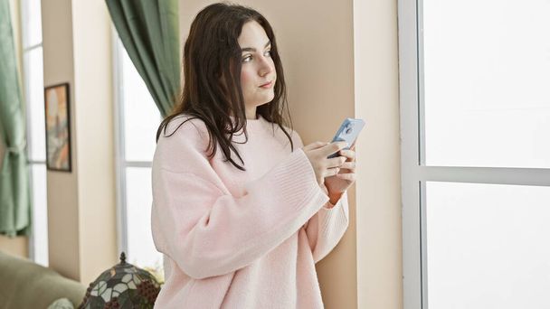 Eine nachdenkliche junge Frau steht zu Hause am Fenster und hält ein Smartphone in einem gemütlichen rosa Pullover - Foto, Bild