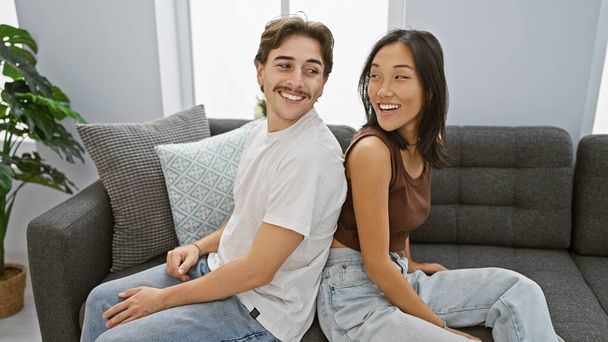 Una pareja interracial se sienta juntos sonriendo en una acogedora sala de estar, encarnando una relación moderna y amorosa en el interior. - Foto, imagen
