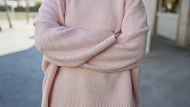 Женщина в розовом свитере стоит со скрещенными руками на городской улице, источая повседневный городской шик. - Фото, изображение