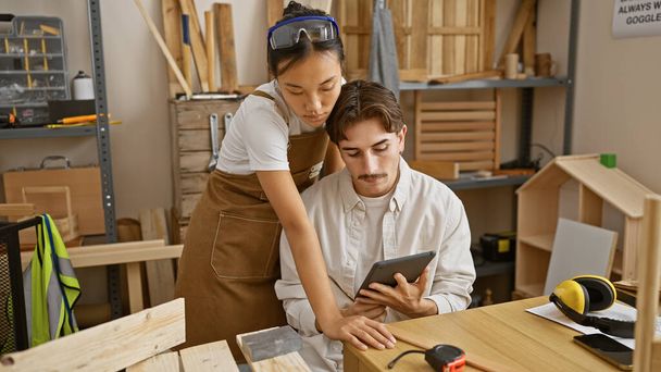 Two carpenters, a man and a woman, collaborate using a tablet in a woodwork workshop surrounded by tools. - Photo, Image