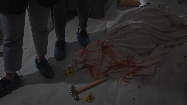Δύο ερευνητές σε καλύμματα παπουτσιών αναλύουν μια αιματηρή σκηνή εγκλήματος με αριθμημένες ετικέτες αποδεικτικών στοιχείων και ένα σφυρί σε εσωτερικούς χώρους. - Φωτογραφία, εικόνα